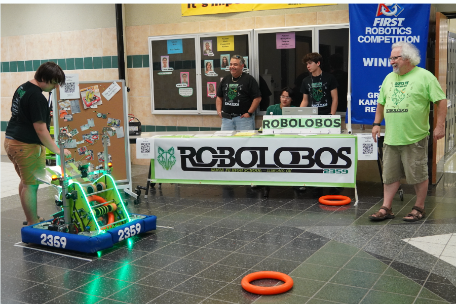 Robotics Club sponsors stand next to their booth, watching as a club member demonstrate one of the club's gadgets. 