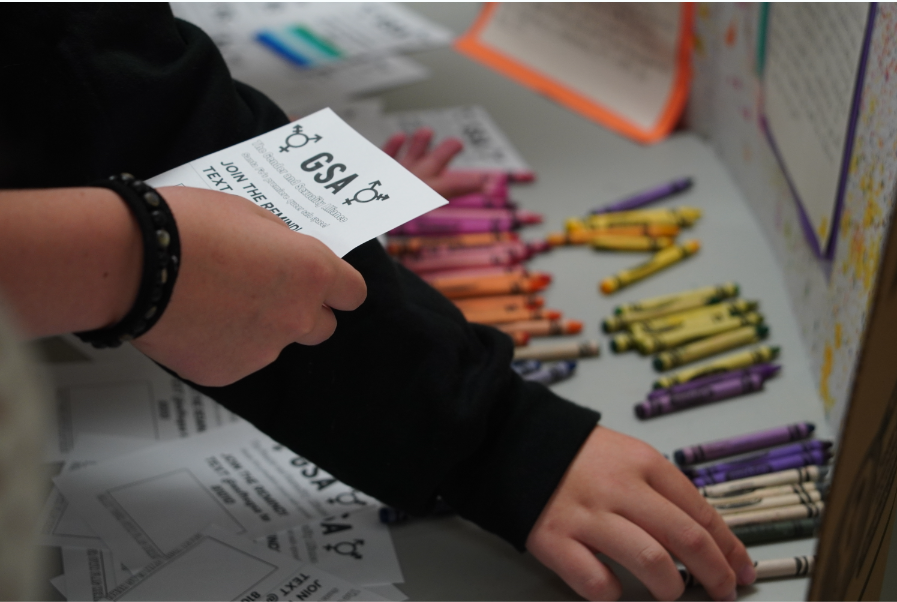 Student holds a GSA pamphlet while another student reaches for a crayon placed on the table to draw attention to the clubs impact.